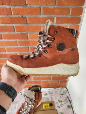 Bruna mockaboots från Rieker 40 - Säljer ett par snygga bruna boots från Rieker i mocka med grov sula och färgglada stickade detaljer vid skaftet. Skorna har mönstrade snören, dragkedja på sidan och mjukt ullfoder för extra värme. Perfekta för höst och vinter!