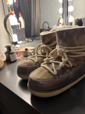 Camouflage moon boots från Inuikii - Säljer ett par moon boots från Inuikii med camouflage-mönster i gröna och bruna toner. Skorna har snörning med grova rep, platt sula och insida i mjuk syntetpäls. Utsidan är i mocka och skinn, perfekt för kalla vinterdagar. säljer då de är för små!