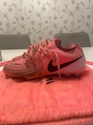 Nike Phantom GX 2 elite rosa fotbollsskor - Nike Phantom GX 2 elite fotbollsskor i en snygg rosa färg med svarta detaljer och logga. Skorna har snörning och en stickad ovandel som ger bra passform. Sulan är platt med dobbar för gräsplan. Kommer med matchande rosa påse. Perfekt för dig som vill sticka ut på planen. Inga skador, endast smutsiga, men går nog att tvätta bort.
