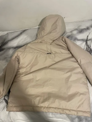 Beige Nike pufferjacka Therma-Fit - Säljer en beige pufferjacka från Nike med Therma-Fit teknologi. Jackan har huva, dragkedja framtill och två sidofickor. Insidan är fodrad med mjuk fleece för extra värme. Perfekt för kalla dagar och har en clean look med diskret Nike-logga på bröstet.