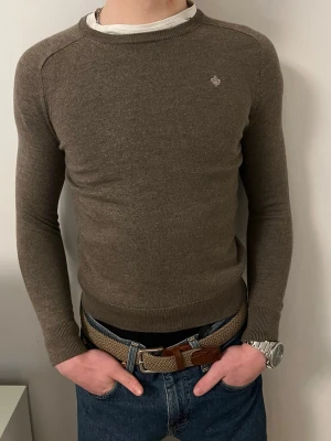 Brun stickad tröja från Morris - ‼️Modellen är 180‼️Snygg brun stickad tröja i merinoull från Morris med rund hals och diskret logga broderad på bröstet. Tröjan har en slim passform och långa ärmar med ribbade muddar. Perfekt att matcha med jeans för en clean och stilren look. 🌟