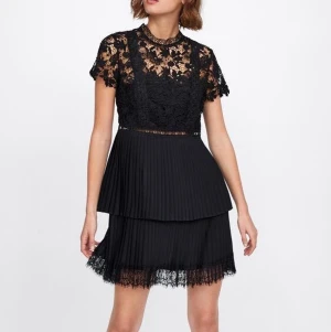 Svart spetsklänning från Zara - Supersnygg svart kortklänning från Zara med blommig spets upptill och korta ärmar. Kjolen är plisserad i flera lager och avslutas med spetsdetaljer. Hög hals och dragkedja bak. Perfekt för dig som vill ha en elegant look med coola detaljer. 