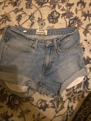 Blå jeansshorts från Acne Studios - Säljer ett par klassiska blå jeansshorts från Acne Studios, modell Max Lit Vintage. De har hög midja, fem fickor, uppvikta ben och råa kanter. Materialet är 100% bomull och de har knapp och dragkedja framtill. Perfekta för sommarens alla dagar!