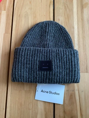 Grå ribbad ull mössa från Acne Studios - Säljer en snygg grå mössa från Acne Studios med ribbad stickning och ett svart patch med ansikte framtill. Mössan är tillverkad i mjuk ull och har en bred uppvikt kant. Perfekt för dig som gillar stilrena accessoarer med en twist. 100% Ull