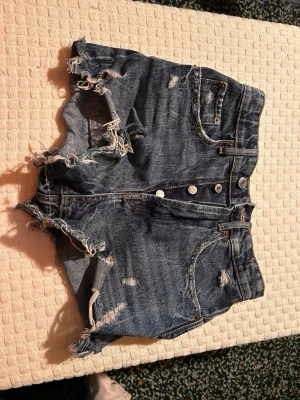 Blå denimshorts från Zara, strl 34 - Säljer ett par blå denimshorts från Zara i storlek 34. De är högmidjade med fransiga och slitna kanter, klassiska fem fickor och knappgylf. Perfekta för sommarens alla äventyr och har en cool, edgy vibe.