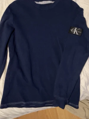  Calvin Klein Jeans - Mörkblå stickad tröja från Calvin Klein Jeans med stort CK-märke på ärmen. Knappt använd. 