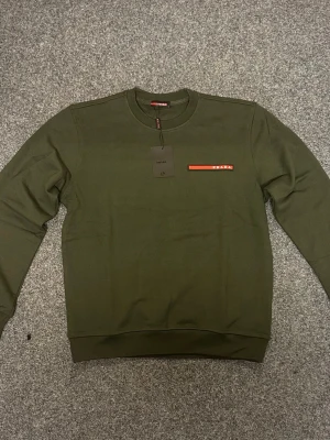 Grön sweatshirt från Prada - Snygg grön sweatshirt från Prada med rund hals och diskret röd logga på bröstet. Tröjan har långa ärmar och ribbade muddar vid ärmslut och nederkant. Perfekt för dig som gillar stilrena designer och exklusiva märken.