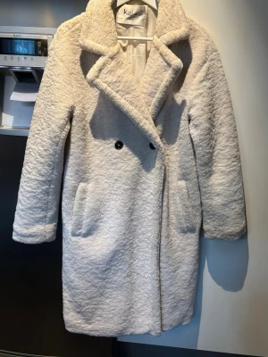 Beige teddykappa från K.Zell Paris - Säljer en beige teddykappa från K.Zell Paris i storlek M. Jackan har bred krage, två svarta knappar framtill och stora fickor på sidorna. Materialet är mjukt och fluffigt, perfekt för kyliga dagar. Jackan har en loose passform och är helfärgad utan mönster.