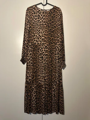 H&M leopardmönstrad klänning, storlek S - Leopardmönstrad klänning med lång ärm och resår vid handlederna. Klänningen har en rund halsringning och en liten öppning med knapp i nacken. Den har en avslappnad passform med en avskärning i midjan och en volangkjol.
