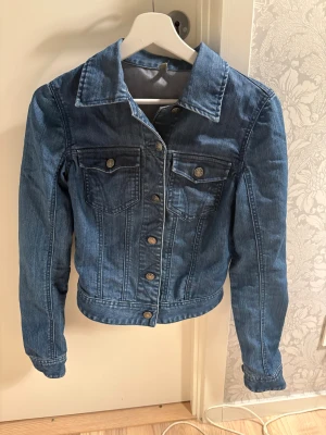 Blå jeansjacka från Vila, storlek S - Snygg klassisk blå jeansjacka från Vila i storlek S. Jackan har krage, två bröstfickor med knappar och långa ärmar. Stängs med knappar framtill och har en normal passform. Perfekt att slänga över en hoodie eller t-shirt för en chill look.