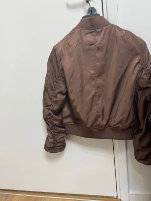 Brun bomberjacka från FB Sister XS - Snygg brun bomberjacka från FB Sister i storlek XS. Jackan har glansigt material, ribbade muddar, dragkedja framtill och coola detaljer som ficka med dragkedja på ärmen. Perfekt för dig som vill ha en trendig och stilren look.