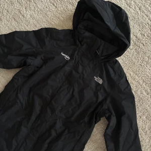 Svart vindjacka från The North Face - Svart vindjacka med huva från The North Face, med logga på bröstet och 'hembla' tryckt på andra sidan. Jackan har dragkedja framtill och är tillverkad i lätt polyester, perfekt för blåsiga dagar. Snygg och enkel design med långa ärmar och praktiska fickor.