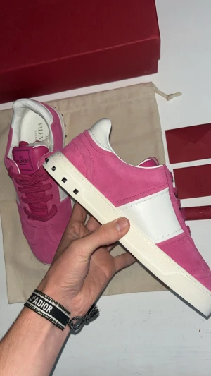 Valentino Garavani Open rosa sneakers 38 - Snygga Valentino Garavani Open sneakers i rosa mocka med vita skinnpaneler och ikoniska nitar på sulan. Skorna har rund tå, platt sula och klassisk snörning. Perfekt statement-piece för dig som vill sticka ut med färg och lyxig känsla.