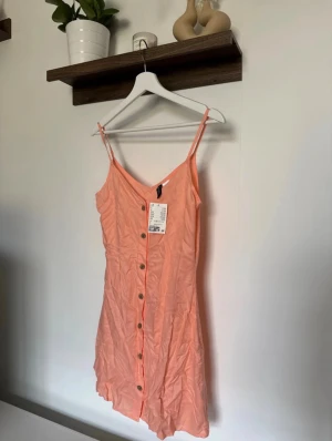 Persikofärgad klänning H&M  - Säljer en persikofärgad kort klänning från H&M i storlek 34. Klänningen har smala axelband, v-ringning och knappar framtill. Tillverkad i viskos. Helt oanvänd med prislapp kvar. 