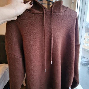 Brun stickad hoodie med huva - Mysig brun stickad hoodie med huva och snörning. Tröjan har långa ärmar och ribbad nederkant, perfekt för chill dagar. Färgen är djup chokladbrun och materialet känns mjukt och skönt mot huden. Enkel och stilren design utan mönster eller logga.