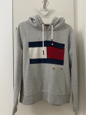 Grå hoodie Tommy Hilfiger Denim XL - Snygg grå hoodie från Tommy Hilfiger Denim i storlek XL. Tröjan har klassisk Hilfiger-logga i blått, vitt och rött framtill, snörning vid huvan och ribbade muddar. Tillverkad i mjuk bomull och polyesterblandning, perfekt för chill dagar.