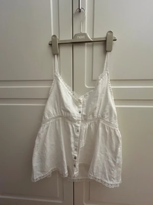 Vit linnetopp med spets och knappar - Söt vit linnetopp från Ambar med smala axelband, spetsdetaljer längs kanterna och knappar framtill. Toppen har en luftig passform och är dekorerad med spets både vid bysten och nederkanten. Perfekt för varma dagar och en fräsch look.