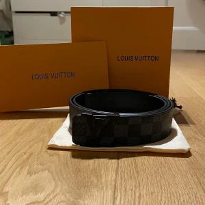  Louis Vuitton Skärp - Snyggt svart Louis Vuitton bälte med klassiskt rutigt mönster och LV-logga på spännet. Bältet är tillverkat i exklusivt skinn och har en stilren, matt finish. Perfekt accessoar för att lyfta din outfit med en lyxig touch. Längd 120 cm