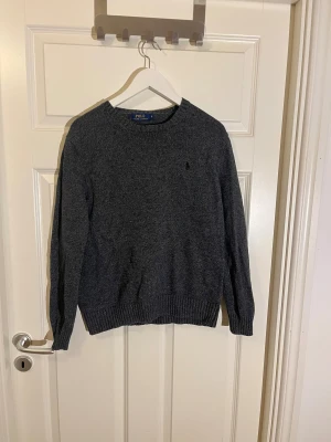 Mörkgrå stickad tröja Polo Ralph Lauren - Mörkgrå stickad tröja från Polo Ralph Lauren i storlek S. Klassisk rund hals och ribbade muddar vid ärmslut och nederkant. Liten broderad logga på bröstet. Perfekt för dig som gillar stilrena och tidlösa plagg. Vid fler frågor skriv// Almen