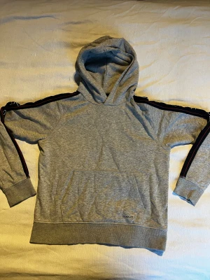 GANT HOODIE - Snygg grå hoodie från GANT med huva och känguruficka. Svarta band med GANT-logga och röda detaljer längs ärmarna ger en sportig vibe. Mjuk bomullsblandning, ribbade muddar och klassisk passform. Perfekt för chill dagar. Använd ett fåtal gånger. Storlek 176/XS.