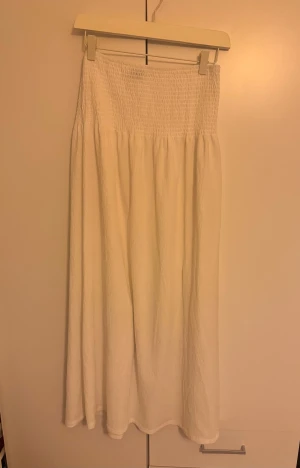 Vit långkjol med smockad midja - Säljer en vit långkjol med smockad midja som ger en skön och flexibel passform. Kjolen är gjord i ett lätt och luftigt material, perfekt för varma dagar. Den har en enkel och stilren design utan mönster eller extra detaljer.