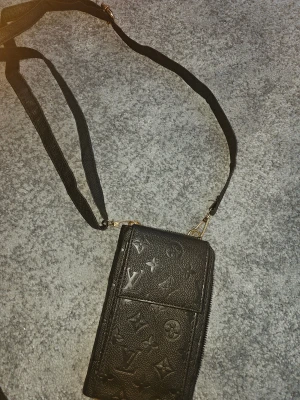 svart axelväska i skinn - Svart axelväska i präglat skinn med klassiskt monogrammönster. Väskan har justerbar axelrem, guldfärgade detaljer och flera fack med dragkedja för kort och småsaker. Perfekt storlek för mobilen och det viktigaste.