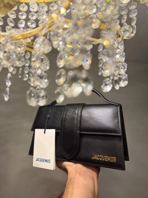 Jacquemus the large bambino - Snygg svart handväska från Jacquemus med guldfärgad logga och detaljer. Väskan har ett stilrent, rektangulärt format med topphandtag och avtagbar axelrem med guldfärgade spännen. Perfekt för dig som gillar design och lyxiga accessoarer.