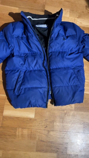 Blå pufferjacka från Calvin Klein Jeans - Säljer en blå pufferjacka från Calvin Klein Jeans med CK-logga på ärmen. Jackan har hög krage, två sidofickor och grov dragkedja framtill. Perfekt för kalla dagar och har en riktigt snygg streetstil. Fodrad insida i svart och coolt märke på insidan.