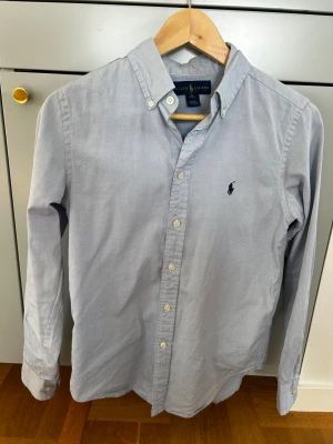 Ljusblå skjorta från Ralph Lauren S - Snygg ljusblå skjorta från Ralph Lauren med klassisk button-down krage och broderad logga på bröstet. Skjortan har långa ärmar och vita knappar framtill. Perfekt för dig som gillar stilrena och tidlösa plagg. 