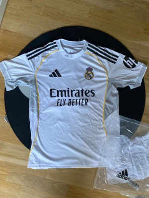 Real Madrid Mbappé tröja Adidas M -  Real Madrid matchtröja med Mbappé och nummer 10 på ryggen. Tröjan är vit med svarta och gula detaljer, korta ärmar och klassiska Adidas-ränder på axlarna. Materialet är lätt polyester, perfekt för fotboll och sport. OBS! Denna tröja är en replika alltså ej autentisk men väldigt bra kvalite!