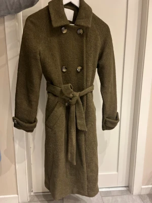 Olivgrön kappa från Samsa Collection - Olivgrön kappa med dubbelknäppning och bälte i midjan från Samsa Collection. Långärmad med klassisk krage och snygga knappar på ärmsluten. Yttermaterialet är en mix av polyester, ull och viskos som ger en mjuk och varm känsla. Kappan är i storlek 34, men jag tycker den känns mer som 36/38. Fint skick!