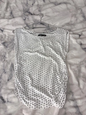 Vit prickig topp från Sinsay XS - Vit ärmlös topp från Sinsay i storlek XS med svarta prickar och snygg rynkdetalj på sidan. Toppen har rund halsringning och är gjord i mjukt bomullsmaterial. Perfekt för dig som gillar en lekfull och trendig stil.