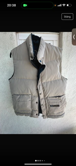 Beige dunväst från Canada Goose - Säljer en beige dunväst från Canada Goose med hög krage och stora fickor framtill. Västen har svarta detaljer på insidan och stängs med både dragkedja och knappar. Perfekt för lager-på-lager under höst och vinter. Storlek S herr 