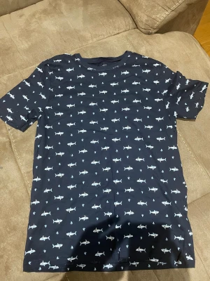 Mörkblå t-shirt med hajmönster - Säljer en mörkblå t-shirt från Old Navy med coolt hajmönster över hela plagget. T-shirten har rund hals och korta ärmar, perfekt för dig som gillar unika prints och vill sticka ut lite extra. Materialet är mjuk bomull som känns skönt mot huden.