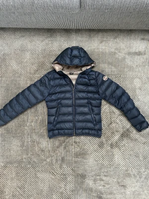 Mörkblå Colmar dunjacka  - Snygg mörkblå dunjacka från Colmar med beige insida och huva. Jackan har ett quiltat mönster, dragkedja framtill och två sidofickor. Märkespatch på ärmen och diskret logga bak. Perfekt för kalla dagar och riktigt stilren look.