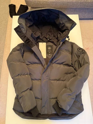 Svart pufferjacka från Canada Goose - Säljer en svart pufferjacka från Canada Goose med stor huva och snyggt broderat märke på ärmen. Jackan har quiltad design, ribbade muddar och dragkedja framtill. Perfekt för kalla vinterdagar och har en clean, modern look.