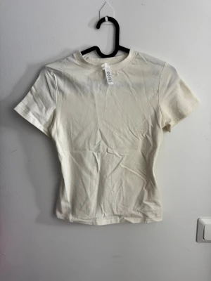 Vit t-shirt från SKIMS i bomull storlek XS - En stilren vit t-shirt från SKIMS i mjuk bomull. Modellen har rund hals och korta ärmar, perfekt för en clean och enkel look. Passformen är normal och tyget är slätt utan mönster eller detaljer. Enkel att matcha med allt i garderoben.