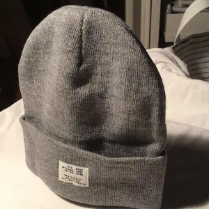 Grå mössa med etikett från Bark - Snygg grå mössa från Bark med uppvikt kant och en vit etikett framtill med text. Enkel och stilren design, perfekt för dig som gillar cleana accessoarer. Materialet är mjukt och stickat, vilket gör den skön att bära under kyliga dagar.