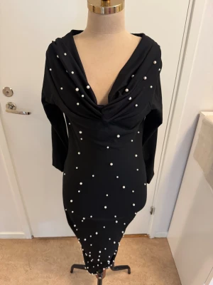 Svart långklänning med pärldetaljer - Svart långklänning med snygg draperad v-ringning och långa ärmar. Klänningen är dekorerad med vita pärlor över hela framsidan för en extra cool look. Tillverkad i stretchigt material av polyester och elastan som sitter skönt på kroppen.