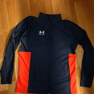 Svart och röd /hoodie  Under Armour - Sportig hoddie  från Under Armour i svart med röda paneler på sidorna. Jackan har hög krage, hel dragkedja framtill och långärmad design. Materialet är polyester och loggan syns tydligt på bröstet. Perfekt för träning eller chill. Storlek M passar smal också riktigt fint passar det mesta funkar som en jacka också med tröja under ..i god skick 