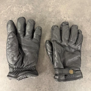 Svarta Hestra handskar i älgskinn - Säljer ett par svarta handskar från Hestra i mjukt älgskinn med isolering av Primaloft. Insidan är fodrad med polyester och mudden är i en mix av ull och polyamid. Handskarna har en snygg detalj med mässingsfärgad knapp där det står Hestra. Storlek 10