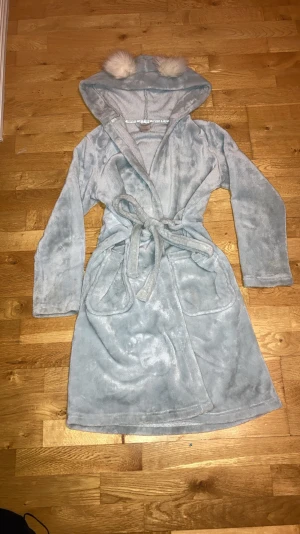 Ljusblå teddyjacka med huva och bollar - Supermysig ljusblå teddyjacka med huva och två fluffiga vita bollar på toppen. Jackan har långa ärmar, stora fickor och knyts med bälte i midjan. Materialet är mjukt fleece som känns skönt mot huden, perfekt för chilliga dagar hemma eller efter duschen. Storlek är 148/152 men passar alla storlekar. Går ner till knäna på en som är 165 lång. PRISET GÅR ATT DISKUTERA (GÅR GÄRNA NER PÅ PRISET VID KÖP)💋