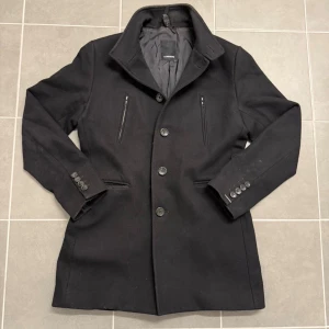 Svart rock från J.Lindeberg - Stilren svart rock från J.Lindeberg med klassisk krage och fem knappar framtill. Jackan har två bröstfickor med dragkedja, två sidofickor och dekorativa knappar vid ärmsluten. Baktill finns en snygg detalj med knappslå. Perfekt för dig som gillar clean och tidlös look. Storlek 46 sitter som M.