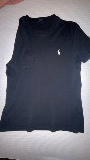 Svart t-shirt från Polo Ralph Lauren - Svart t-shirt från Polo Ralph Lauren med klassisk rund hals och vit broderad logga på bröstet. Enkel och stilren design i mjuk bomull, perfekt för en clean och avslappnad look.