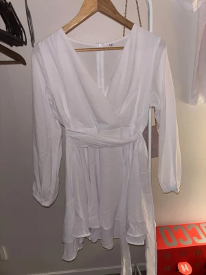 Vit omlottklänning med volang - Superfin vit omlottklänning med långa ärmar och volangdetalj nertill. Klänningen har v-ringning och knyts i midjan med ett bälte för en snygg passform. Perfekt för sommarens alla tillfällen!💕