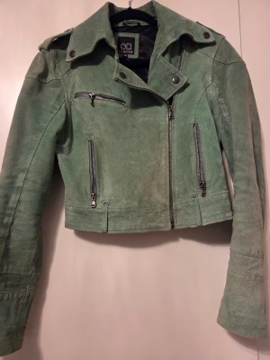 Grön mocka bikerjacka från H Collection - Snygg grön bikerjacka i mocka från H Collection. Jackan har croppad passform, coola dragkedjor framtill och på fickorna samt stora slag med tryckknappar. Perfekt för dig som vill sticka ut med en unik jacka i garderoben.