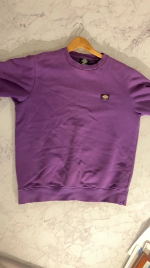 Lila sweatshirt från Dickies - Säljer en lila sweatshirt från Dickies med klassisk rund hals och liten Dickies-logga på bröstet. Tröjan har ribbade muddar vid ärmslut och nederkant. Perfekt för dig som vill ha en färgklick i garderoben och gillar streetwear vibes.