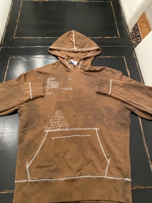 Brun hoodie från COLLUSION med tryck - Snygg brun hoodie från COLLUSION med vita kontrastsömmar och tryck på bröstet och fickan. Hoodien har en stor magficka och coolt grafiskt mönster framtill. Materialet är mjukt och skönt bomullstyg. Perfekt för en avslappnad streetstyle-look.