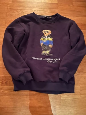 Mörklila Polo Bear sweatshirt s - Säljer en mörklila sweatshirt från Polo Ralph Lauren i storlek M. Framsidan har en stor Polo Bear-tryck med coola detaljer och färger. Tröjan har rund hals, ribbade muddar och är gjord i mjukt bomullsmaterial. Perfekt för dig som gillar streetwear och snygga prints.