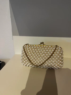 Vit pärlbroderad kuvertväska med kedja - Superfin kuvertväska täckt av vita pärlor och små glittriga stenar, med guldfärgad metallram och matchande kedja. Insidan är fodrad i ljus satin. Perfekt för dig som vill sticka ut med en elegant accessoar.